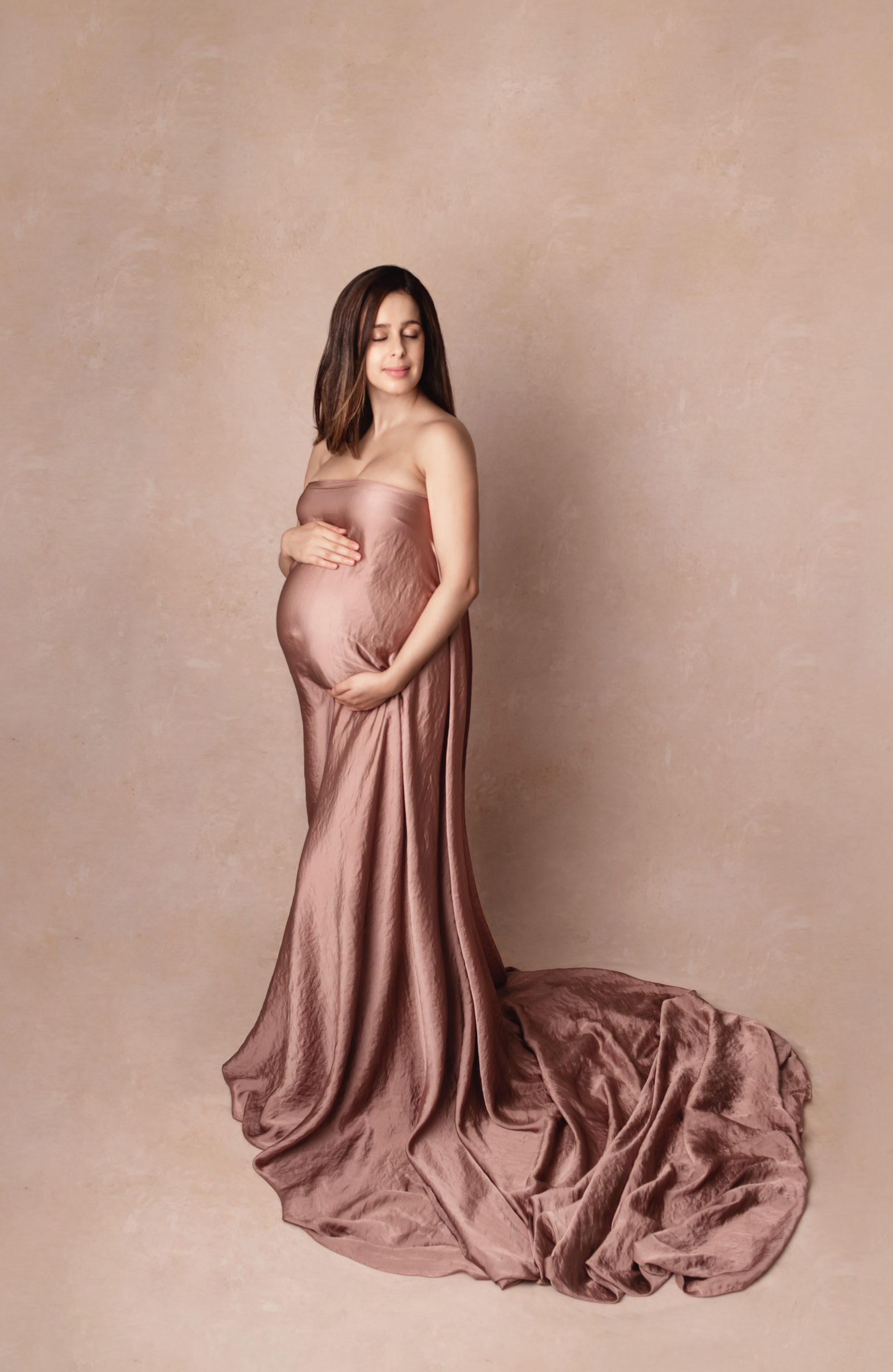 Photo grossesse de belle femme enceinte dans la robe Mii-Estilo avec le font de studio beige réalisée pendent une séance grossesse stylisée avec maquillage et coiffure professionnelles