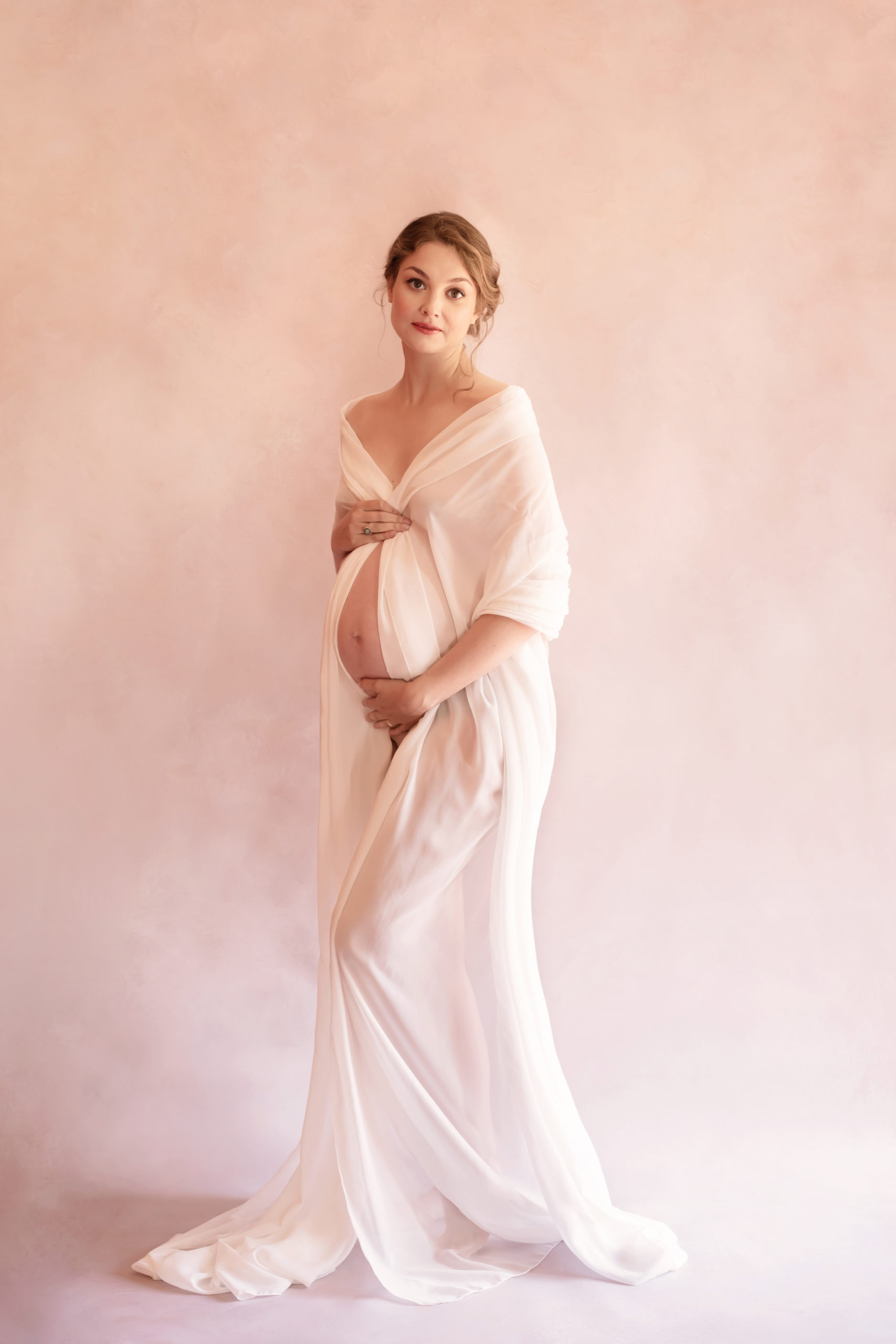 Photo grossesse de belle femme enceinte dans la robe Mii-Estilo avec le fond de studio beige réalisée pendent un shooting grossesse stylisée avec maquillage et coiffure professionnelles